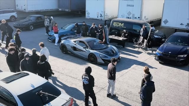 Drone view of a crowd around a grey McLaren with its doors up, trailers parked behind.
